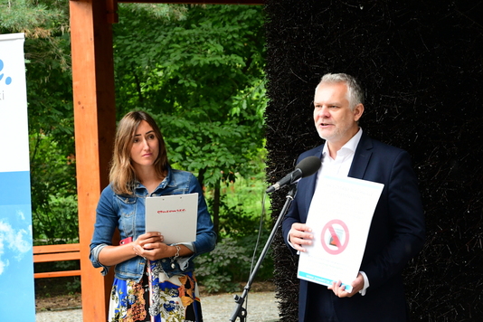 Po lewej stronie kobieta z podkładką i programem konferencji, po prawej stronie mężczyzna mówi przez mikrofon i pokazuje planszę z opisem zmian.