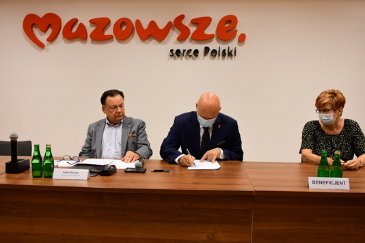 Mężczyźni siedzą przy stole, w tle logo mazowsza. Jeden z nich podpisuje dokumenty