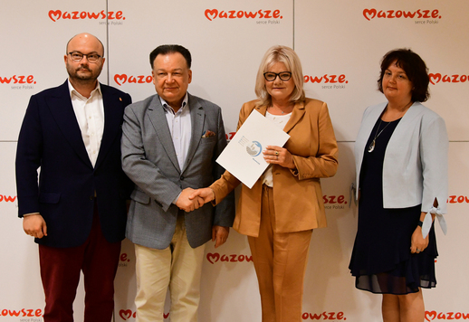 Grupa elegancko ubranych osób stoi na tle logo Mazowsza. Kobieta i mężczyzna pośrodku ściskają swoje dłonie, kobieta prezentuje podpisana umowę