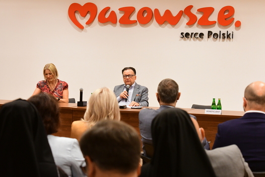 Marszałek siedzi za stołem konferencyjnym. W prawym ręku trzyma mikrofon i przemawia