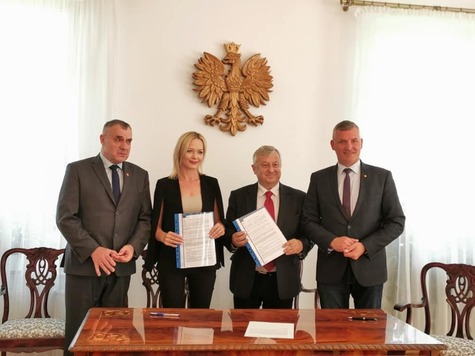 Zdjęcie pamiątkowe. Na tle białej ściany stoi trzech mężczyzn w garniturach oraz elegancko ubrana kobieta. Jeden z mężczyzn oraz kobieta trzymają dokumenty. Na ścianie wisi godło Polski.