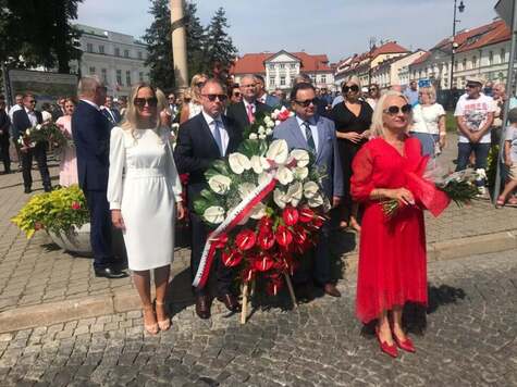 Marszałek z delegacją (mężczyzna w garniturze i dwie kobiety) stoją, trzymając przed sobą wielki wieniec z biało-czerwonych róż