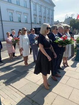 Dwie kobiety i dwóch mężczyzn w garniturach stoją obok siebie. Mężczyzna stojący pośrodku trzyma wieniec z biało-czewonych kwiatów