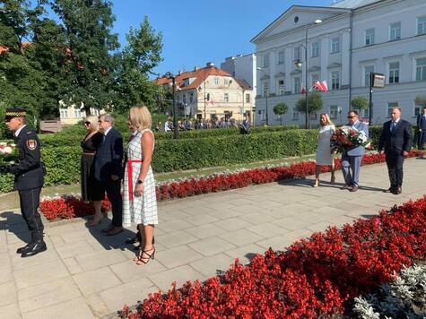 Dwie kobiety i mężczyna stoją obok siebie na szerokim chodniku. Za nimi idzie marszałek z wieńcem, obok niego idzie kobieta