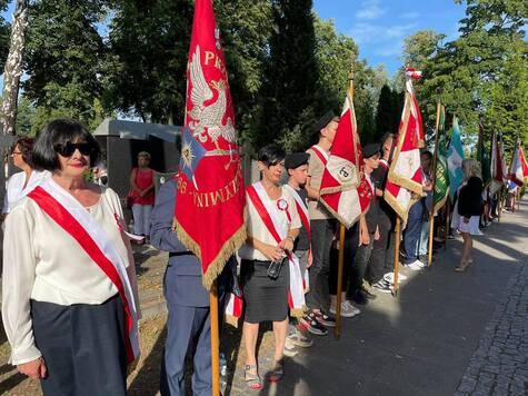 Przedstawiciele organizacji społecznych ze sztandarami stoją wzdłuż głownej alei cmentarza