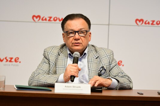 Marszałek siedzi przy stole konferencyjnym, w prawym ręku trzyma mikrofon. Patrzy przed siebie