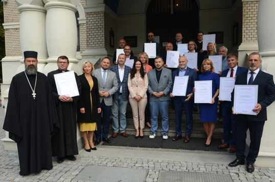 Grupa elegancko ubranych osób stoi na schodach do cerkwi, prezentują certyfikaty 