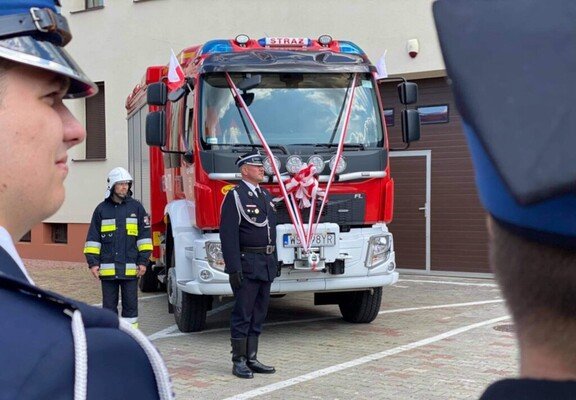 Na głównym planie ozdobiony szarfą wóz strażacki, przed nim stoi druh w mundurze galowym