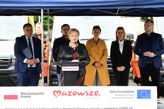 Przemawia kobieta przy mównicy, w tle uczestnicy wydarzenia i karetki