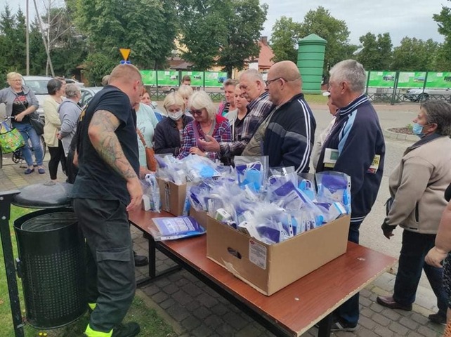 Na stole stoi karton z maseczkami. Po jednej stronie stołu stoi strażak, po drugiej kolejka mieszkańców odbierających pakiety ochronne