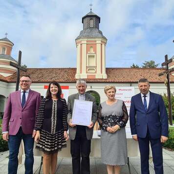 Grupa osób stoi na tle budynku z wieżą, jedna trzyma dyplom