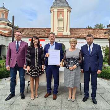 Grupa osób stoi na tle budynku, jeden mężczyzna trzyma dyplom