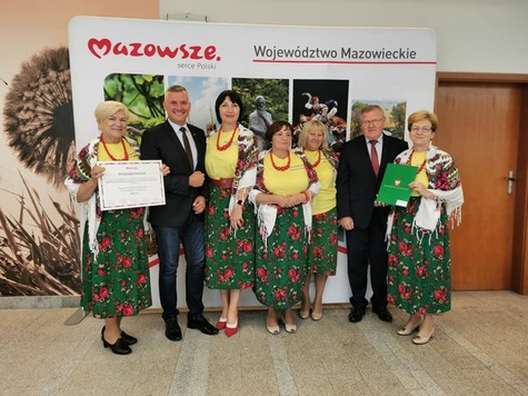 Grupa osób na tle ścianki województwa mazowieckiego. W środku kobiety w strojach ludowych, po bokach mężczyźni w garniturach.