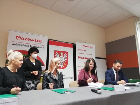 Grup osób za stołem podpisuje umowy, w tle banery promocyjne.