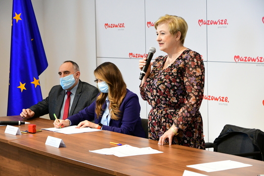 Kobieta stoi przy stole konferencyjnym. W prawej dłoni trzyma mikrofon, do którego przemawia. Po jej prawej stronie siedzi kobieta i mężczyzna w garniturze