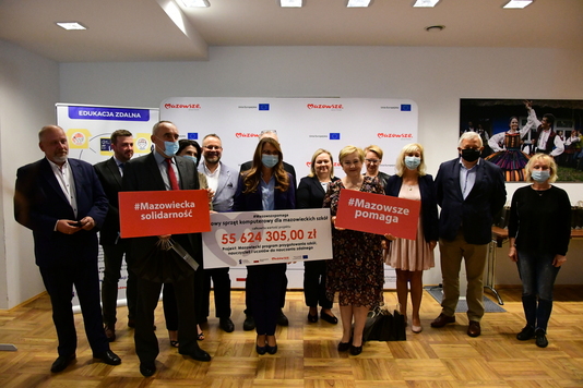 grupa beneficjentów z członek zarządu Elżbietą Lanc oraz wiceprezydent Renatą Kozłowską na przedzie pozuje do zdjęcia. Pani wiceprezydent trzyma wielki czek, Elżbieta Lanc - tablicę Mazowsze pomaga, a mężczyzna stojący obok trzyma tablicę Mazowiecka Solidarność