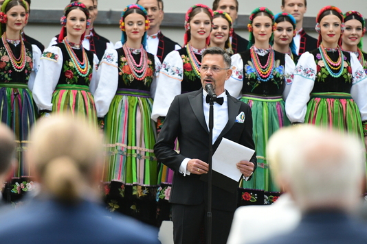 Mężczyzna w garniturze stoi przed mikrofonem i przemawia do publiczności. Głowę ma zwróconą w prawo. Za nim w tle widać stojących w dwóch rzędach członków zespołu mazowsze