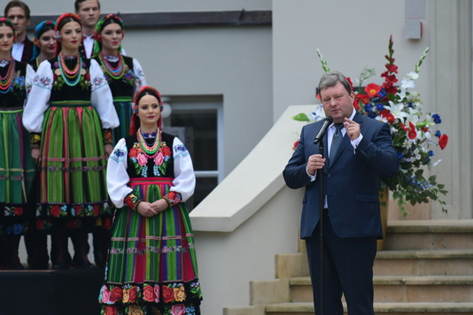 Mężczyzna w garniturze stoi na schodach centrum i przemawia do widzów zgromadzonych przed budynkiem. Obok niego widać tancerkę w stroju ludowym