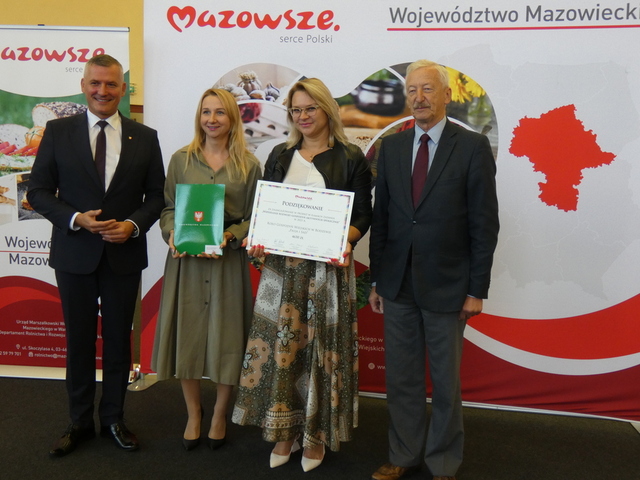 Dwóch mężczyzn i dwie kobiety stoją na tle ścianki promocyjnej Mazowsza. Jedna kobieta trzyma symboliczny czek, druga zieloną teczkę.