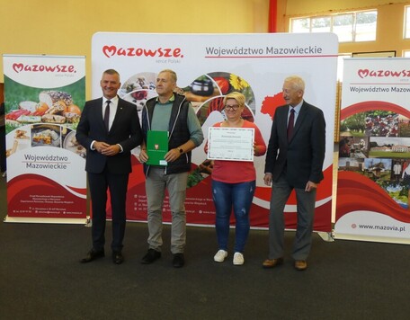 Trzech mężczyzn i kobieta stoją na tle ścianki promocyjnej Mazowsza. Kobieta trzyma symboliczny czek, mężczyzna obok trzyma zieloną teczkę.