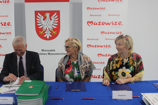 Dwie kobiety z koła gospodyń wiejskich siedzą przy stole konferencyjnym, obok siedzi przedstawiciel urzędu marszałkowskiego - podpisuje umowę