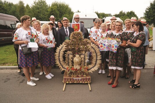 Grupa kobiet w regionalnych strojach i mężczyzn w garniturach stoi przy wielkim wieńcu dożynkowym splecionym z kłosów zbóż