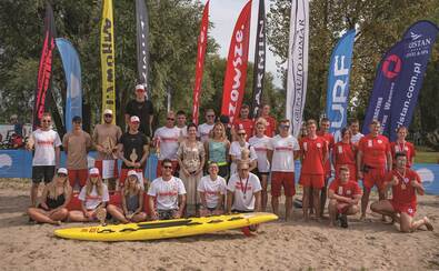 na plaży do zdjęcia pozuje grupa osób, przed nimi kajak, w tle flagi reklamowe