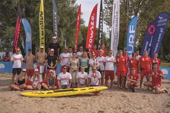 na plaży do zdjęcia pozuje grupa osób, przed nimi kajak, w tle flagi reklamowe