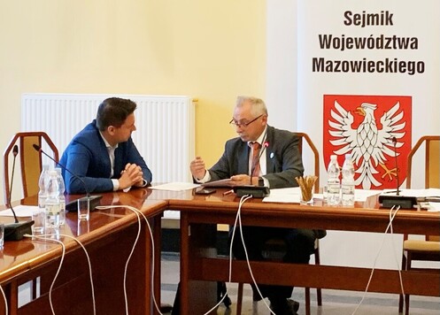 Dwóch radnych w rozmowie, siedzą na przyległych koncach stron stołu.