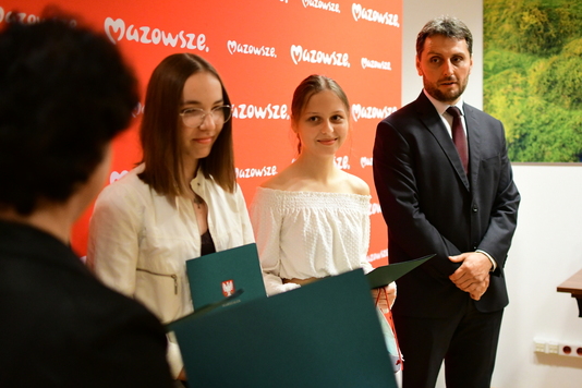 Od lewej na tle  czerwonej ściany z napisem Mazowsze stoją dwie dziewczynki z białych bluzkach oraz mężczyzna w czarnym garniturze