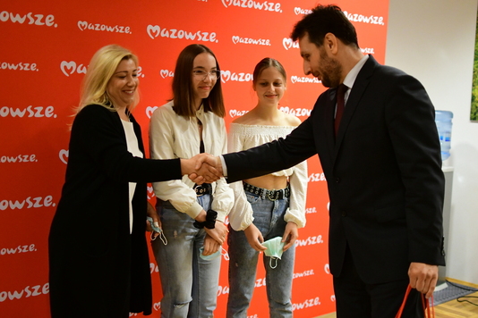 Mężczyzna w czranym  garniturze gratuluje kobiecie w czarnym długim swetrze i blond włosach. Obok niej stoją dwie dziewczynki w białych bluzkach i uśmiechają się