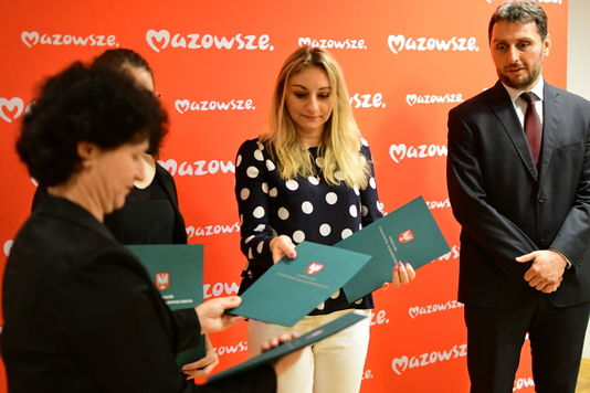 Od lewej stoją kobieta w czarnej marynarce oraz blondynka z długimi włosami w czarnej bluzce w białe grochy. Kobiety  trzymają w dłoniach zielone teczki z orłem