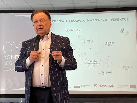 Mężczyzna w marynarce przemawia na tle wyświetlanej prezentacji