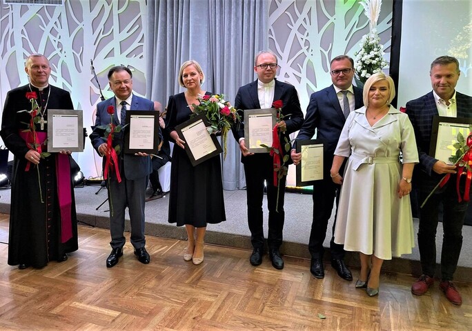 Ksiądz biskup w sutanie, marszałek oraz trzech innych mężczyzn w garniturach i dwie kobiety stoją obok siebie i patrzą na kamerę. Wszyscy trzymają w ręku różę i dyplom. Uśmiechają się.