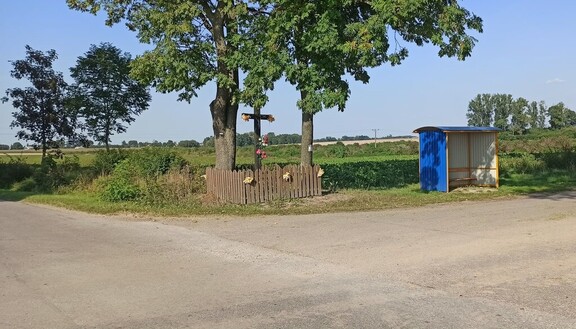 Skrzyżowanie wiejskich dróg. Przy drodze stoi drewniany krzyż otoczony płotem, a obok niego nowa wiata przystankowa. W tle widać łąki i drzewa.