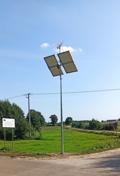 Przy wiejskiej szutrowej drodze stoi wysoki słup na szczycie którego znajdują się panele fotowoltaiczne. Do nich przymocowana jest lampa