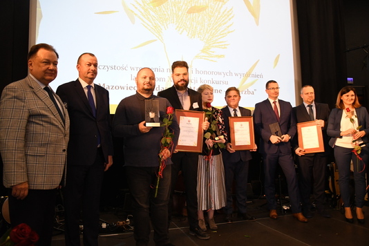 Dziewięć osób stoi na scenie i pozuje do zdjęcia. W rękach trzymają dyplomy i kwiaty