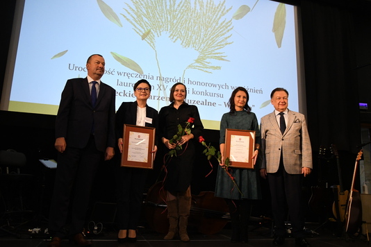 Na scenie stoi pięć osób. Trzy z nich to kobiety laureatki konkursu, które trzymają w rękach nagrody. Z boku po lewej i po prawej stronie stoją dwaj elegancko ubrani mężczyźni