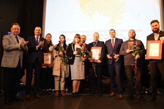 10 elegancko ubranych stoi na scenie. Dwóch mężczyzn z lewej strony bije brawo