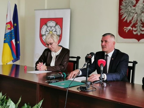 Wicemarszałek Rajkowski siedzi przy stole konferencyjnym i przemawia do mikrofonów ustawionych przed nim na stole. Obok niego siedzi kobieta.