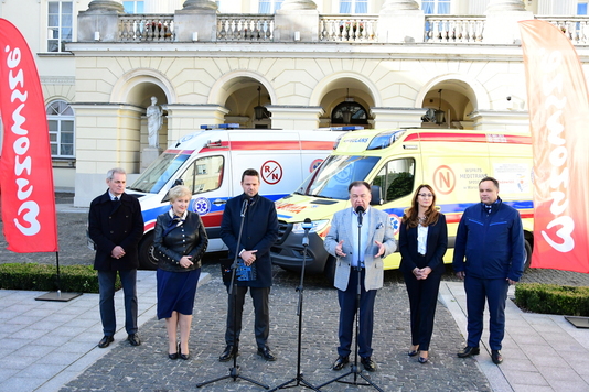 6 osób stoi na tle dwóch karetek, mężczyzna w środku mówi do mikrofonu 