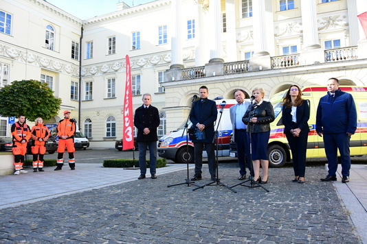 Grupa osób stoi na tle nowoczesnych karetek, kobieta w czarnej, skórzanej kórtce mówi do mikforonu 