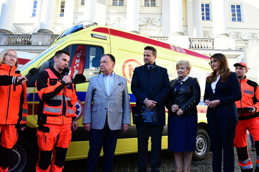 Pięć osób stoi na tle żółtej karetki, ratownik w czerwonym stroju mówi do mikrofonu 