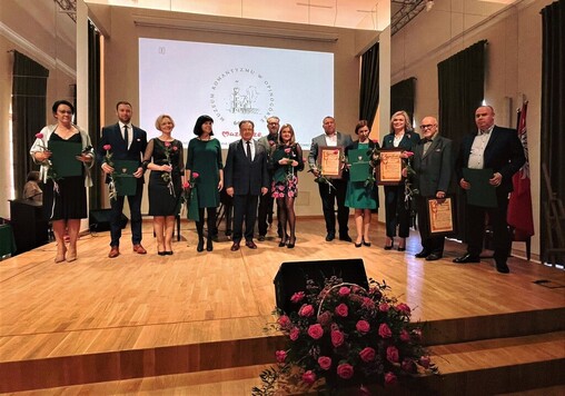 marszałek oraz wyróznieni medalami i dyplomami uznania pracownicy muzeum stoją na scenie i pozują do zdjęcia. Trzy osoby trzymają przed sobą dyplomy. Każdy w ręku ma różę