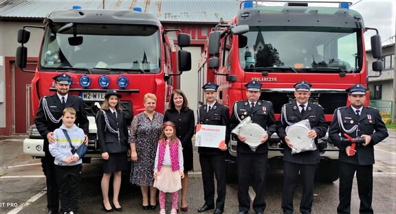 Członek zarządu Orzełowska stoi wraz z kilkoma strazakami w mundurach na tle dwóch wozów strażackich. Przed nimi stoją też dziewczynka i chłopczyk. Strażak stojący obok członek zarządu trzyma tablicę z wypisaną kwotą dofinansowania