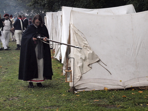 Żołnierz z epoki napoleońskiej w długim płaszczu stoi przed jednym z namiotów ustawionych w rzędzie