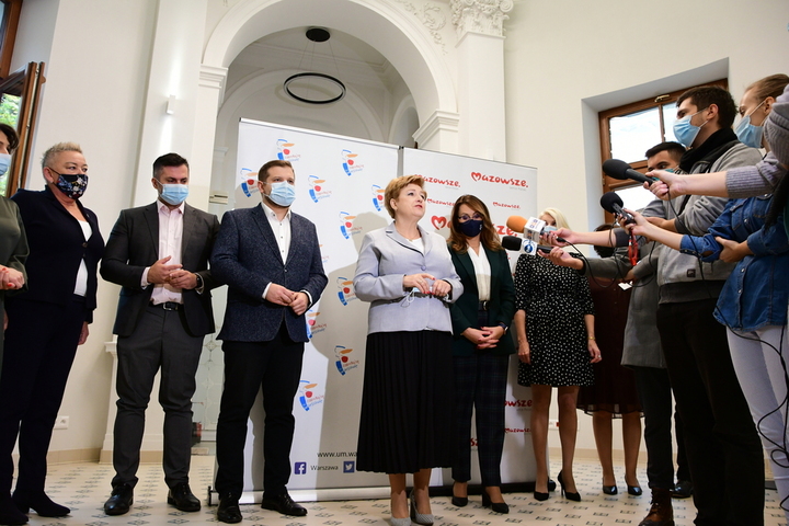 Elżbieta Lanc stoi na środku holu, za nią widać grupę przedstawicieli samorządów uczestniczących w konferencji. Mają maseczki na twarzy