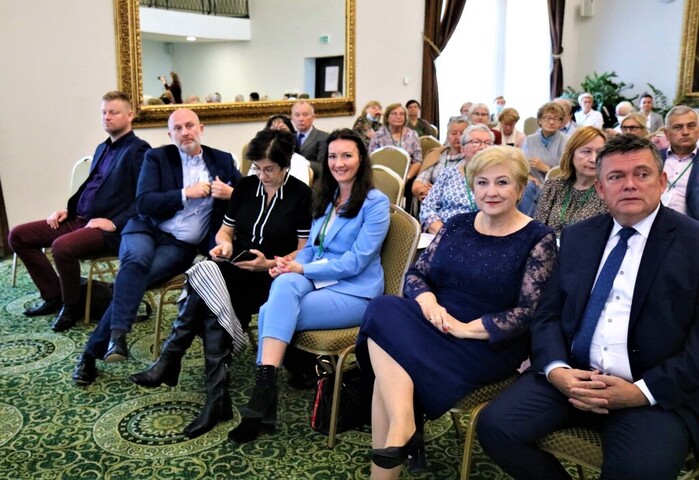 Widok na salę wypełnioną słuchaczami konferencji. Wszyscy siedzą na krzesłach ustawionych w rzędach. W pierwszym rzędzie siedzi Elżbieta Lanc, która patrzy się w kamerę i uśmiecha