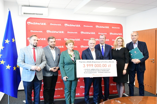 Grupa osób pozuje z czekiem do zdjęcia pamiątkowego na tle ścianki promocyjnej.