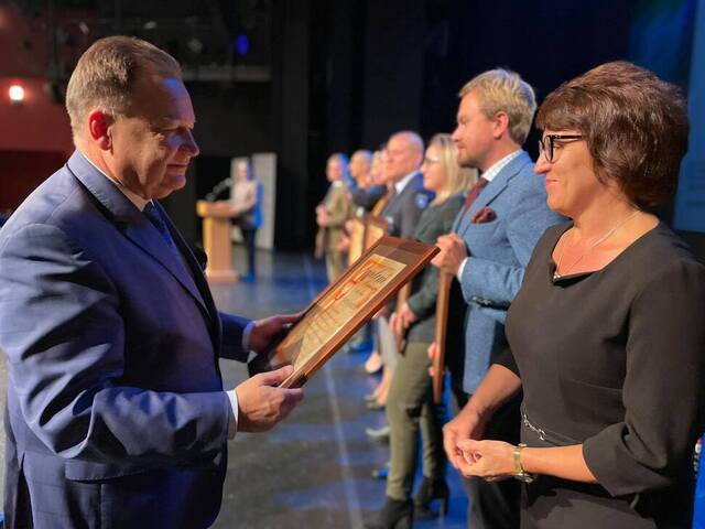 Marszałek stoi bokiem i z uśmiechem przekazuje dyplom kobiecie stojącej na przeciw niego. W tle widać inne osoby, które ustawione w rzędzie za kobietą, czekają na odznaczenie